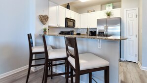 A modern kitchen with white cabinets, a stainless steel refrigerator, and a granite countertop. There are two wooden chairs with white cushions.