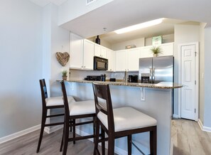 A modern kitchen with white cabinets, a stainless steel refrigerator, and a granite countertop. There are two wooden chairs with white cushions.