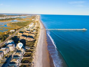 An aerial view of a coastal area with a long pier extending into the ocean, a beach with a wooden walkway, and a row of buildings along the shore.