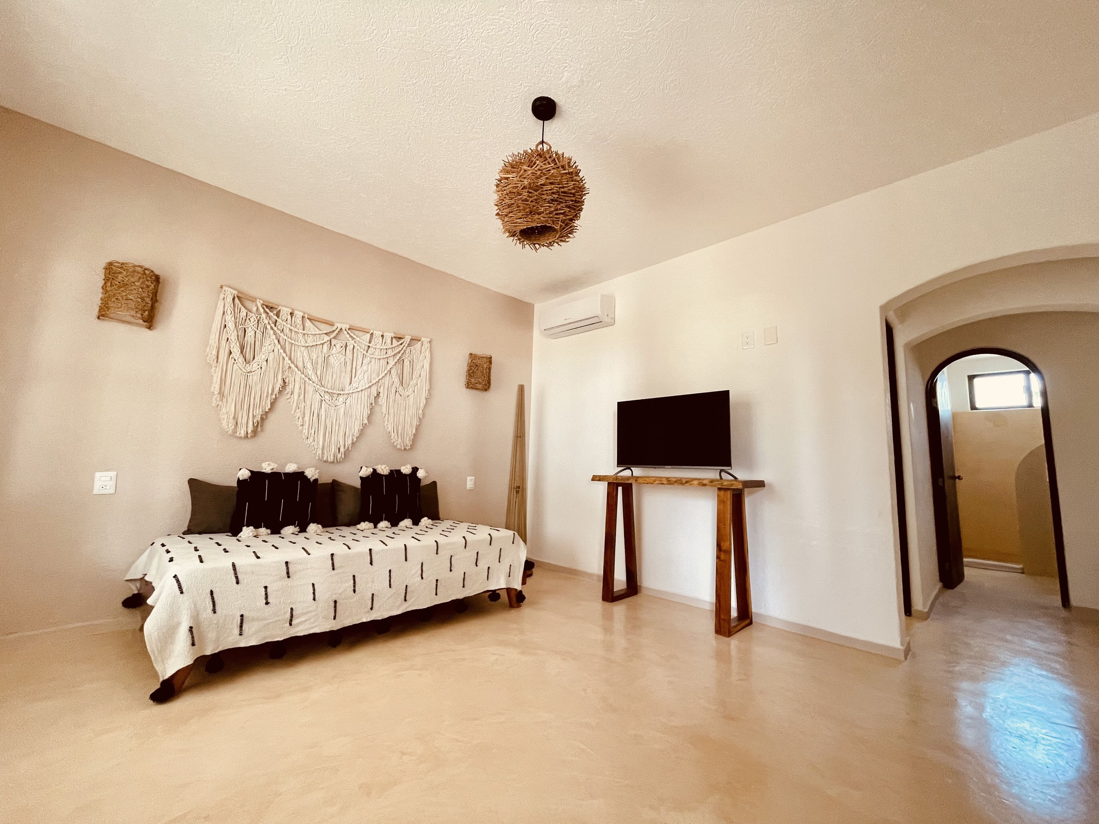 Una habitación amplia con cama, televisor sobre un mueble, un tapiz de macramé y un acceso a otra habitación.