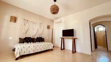 Una habitación amplia con cama, televisor sobre un mueble, un tapiz de macramé y un acceso a otra habitación.
