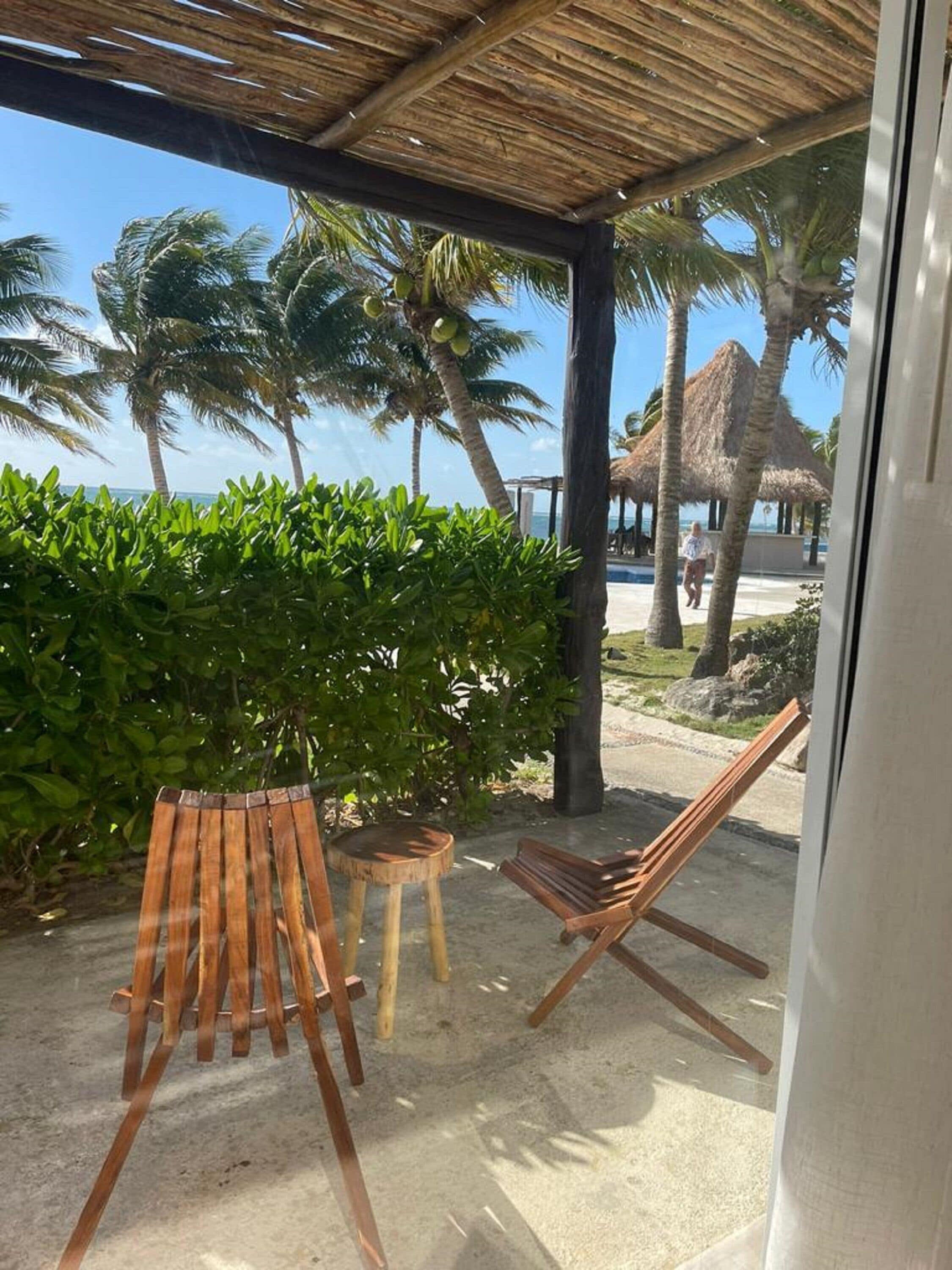 Een overdekt terras met houten stoelen en een klein krukje, met uitzicht op een strand met palmbomen en een rieten gebouw.