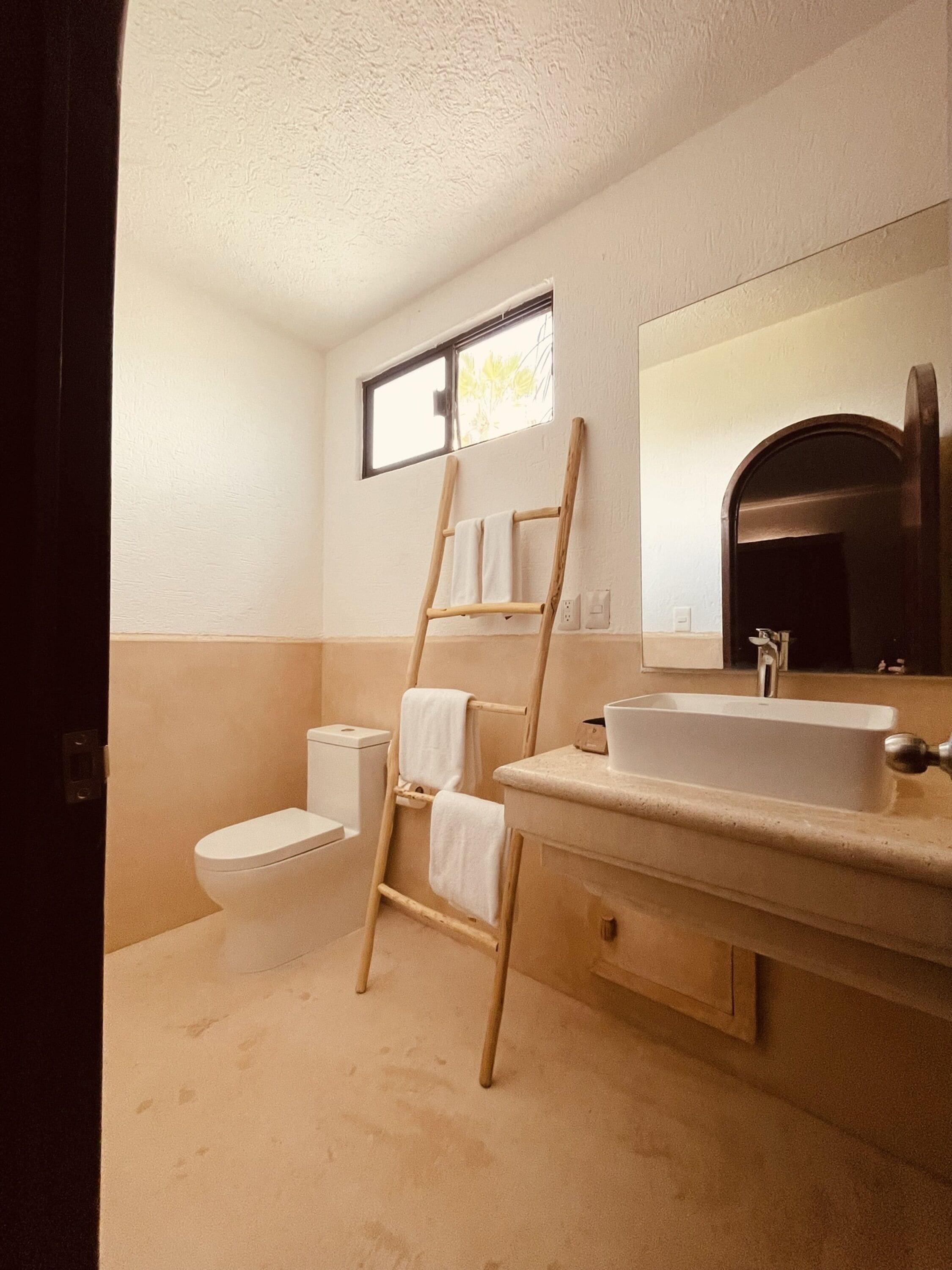 Un baño con inodoro, una escalera de madera, lavamanos y espejo.