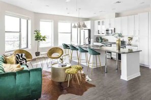 A modern kitchen with white cabinets, a central island, and bar stools. There is a green sofa and a cowhide rug in the living area.