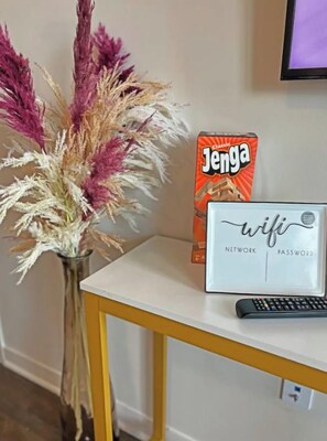 A white vase with dried pampas grass, a box of Jenga, and a white sign with the word "wifi" on a yellow table. - All Things PINK! 1B1B W/Rooftop pool. (Dallas)