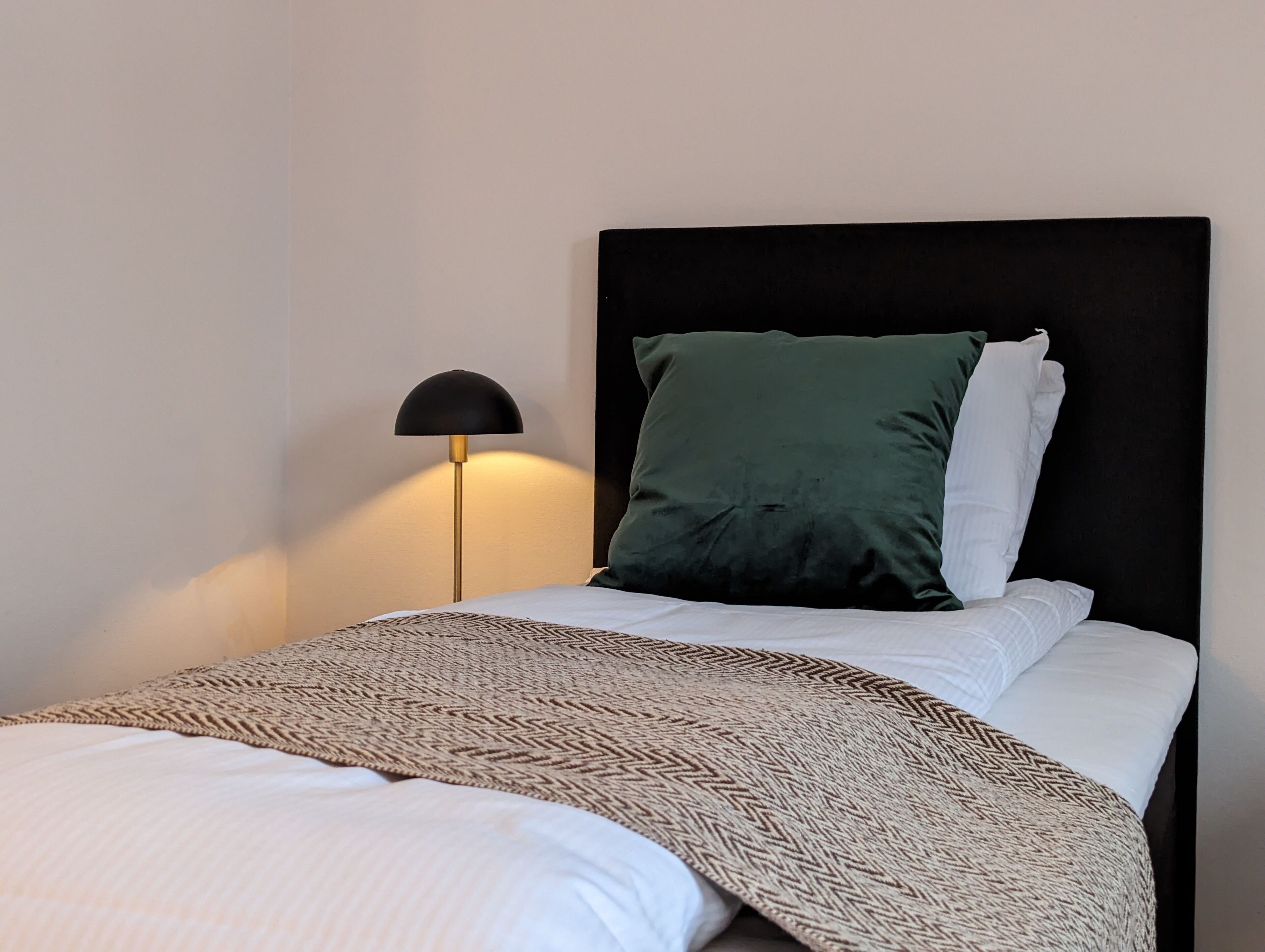 Un lit bien fait, agrémenté d’un couvre-lit à motifs, d’une tête de lit noire et d’une lampe sur pied.