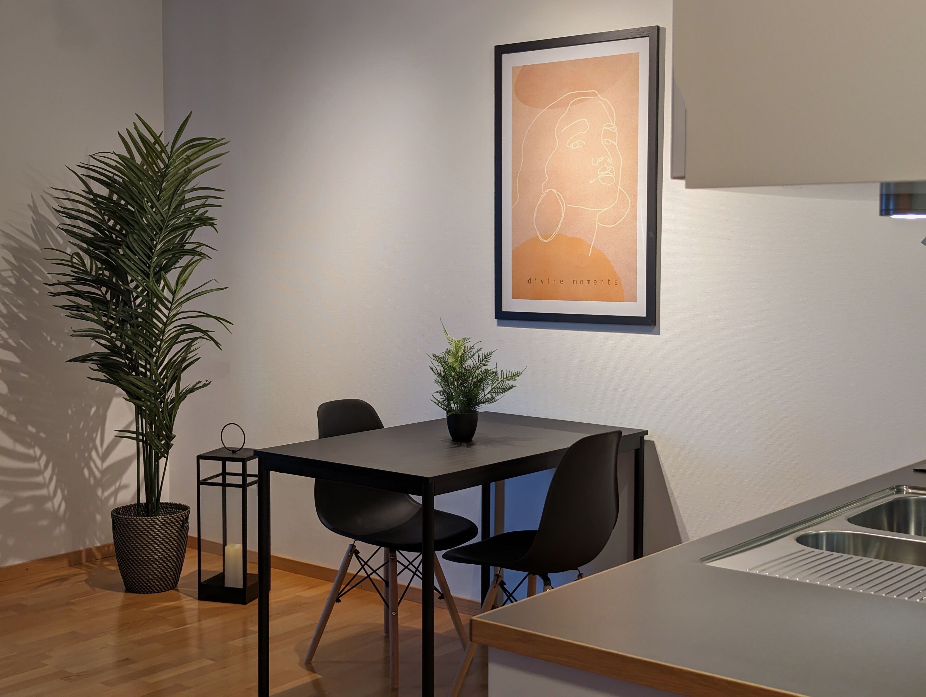 Et moderne køkken med træbordplade, vask og spiseområde med bord og stole. Der er en potteplante på bordet og et indrammet kunstværk på væggen.