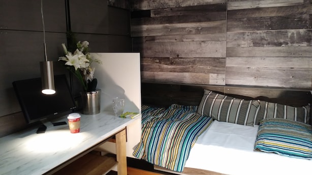 Quarto moderno com cabeceira de madeira, escrivaninha com computador, xícara de café e planta em vaso.