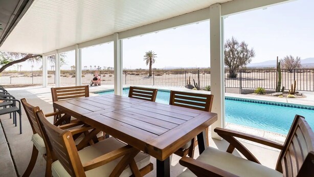 A covered patio with a pool, wooden table and chairs, and a view of the desert landscape.