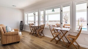 A spacious room with a dining area featuring wooden chairs and a table, large windows with white frames, and a wicker chair with a cushion.