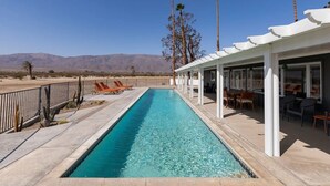 A rectangular pool with clear blue water, surrounded by a white pergola with seating, and palm trees in the background.