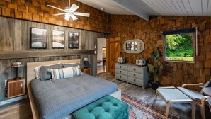 A bedroom with a wooden wall, a bed, a dresser, a chair, and a window with a view of greenery. - Log cabin w/riverfront & hot tub (Rileyville)