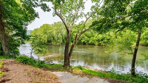 A riverbank with a concrete path leading to a river, surrounded by lush greenery and trees. - Log cabin w/riverfront & hot tub (Rileyville)