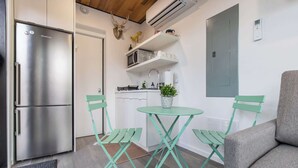 A compact kitchen with a stainless steel refrigerator, a small dining table with two chairs, and a grey sofa.