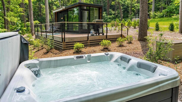 A hot tub in a forested area with a wooden deck and a glass-enclosed cabin in the background.