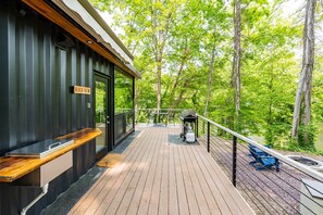 Une terrasse en bois avec un banc et un barbecue, donnant sur une zone boisée.