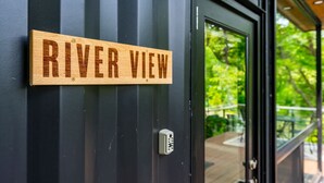 A wooden sign with "RIVER VIEW" on a dark door, a keypad lock, and a view of greenery through a glass door. - Riverside Tiny Cabin w Hot Tub, Fire Pit, & Kayaks (Luray)