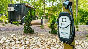 A portable electric vehicle charging station in a natural setting with a small cabin in the background. - Riverside Tiny Cabin w Hot Tub, Fire Pit, & Kayaks (Luray)