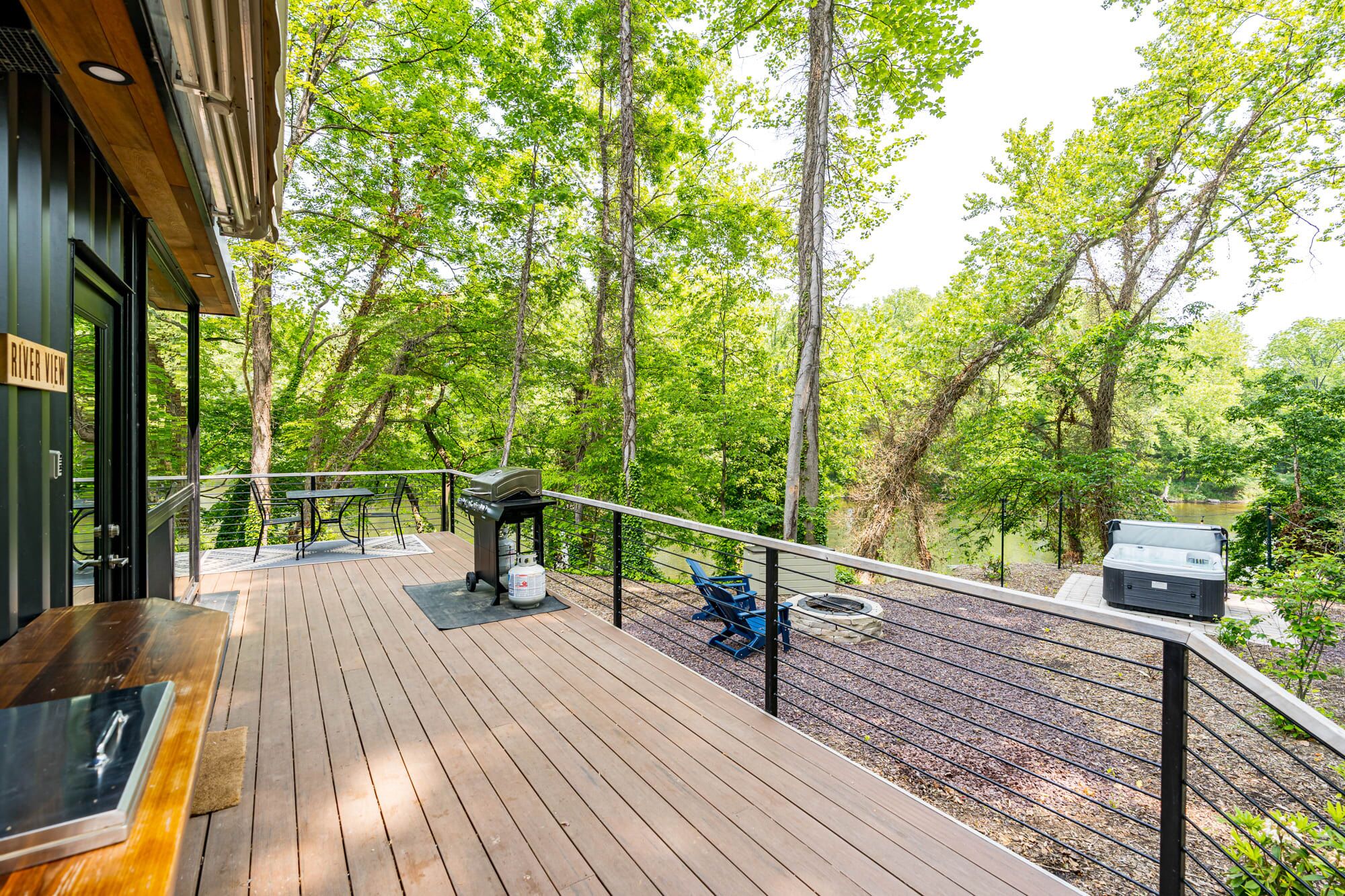 Terraza de madera con una parrilla y zona de estar, rodeada de árboles y con vistas a un cuerpo de agua.
