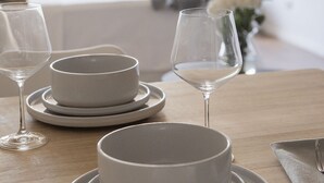 Mesa de jantar com pratos e copos brancos, um vaso com flores brancas e vista para uma varanda com uma planta.