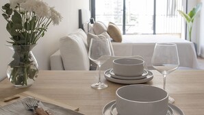 Mesa de jantar posta com pratos, tigelas e copos de vinho brancos, um vaso com flores e uma tábua de corte de madeira com uma faca.