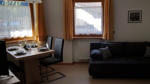 Un salon comprenant un coin repas, un canapé noir, une table dressée pour deux personnes et une fenêtre donnant sur des immeubles.