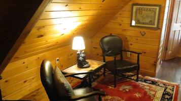 A cozy attic room with wooden walls, a red floral carpet, a table with a lamp, and two leather chairs.