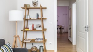 Ein modernes Wohnzimmer mit einem grauen Sofa, einem Holzleiterregal, einer Stehlampe und einem grauen Teppich. Ein Flur führt in einen anderen Raum mit einem Tisch und Stühlen.