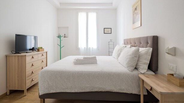 Ein Schlafzimmer mit einem Bett, einem Fernseher auf einer hölzernen Kommode, einem Fenster mit durchsichtigen Vorhängen und einem gerahmten Bild an der Wand.