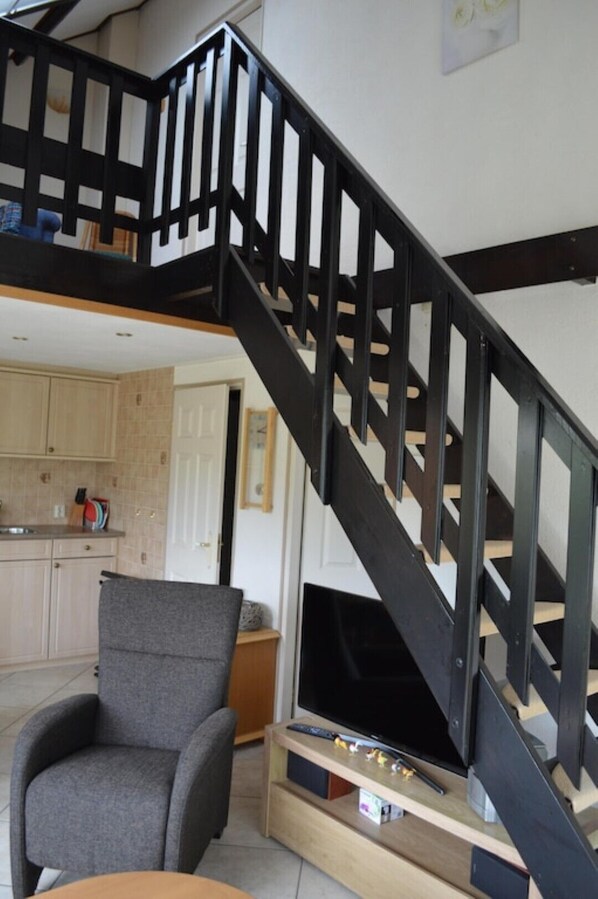 A staircase with black railings leading to an upper level, a grey armchair, a flat-screen TV on a wooden stand, and a kitchen area with cabinets and a sink.