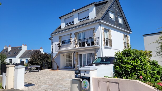 Un immeuble résidentiel de deux étages doté d’un balcon, d’une façade en pierre et d’une allée menant à une voiture garée.