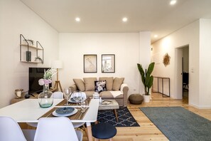 A modern living room with a white sofa, a dining table with chairs, and a coffee table. There is a large plant and a decorative wall piece.