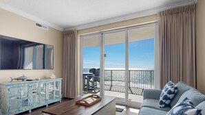 A living room with a large TV, a wooden coffee table, a sofa, and a glass cabinet. There is a balcony with a view of the ocean. - ☀️Beach Service Included-2BR Ocean Villa 803☀️ (Panama City Beach)