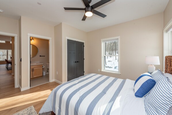 A bedroom with a bed, a ceiling fan, a window with a view of trees, and a door leading to a bathroom.