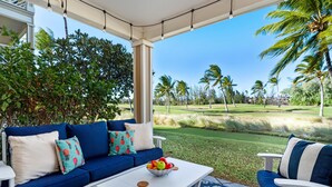 A covered outdoor seating area with a blue sofa, white armchairs, and a coffee table with a fruit bowl.