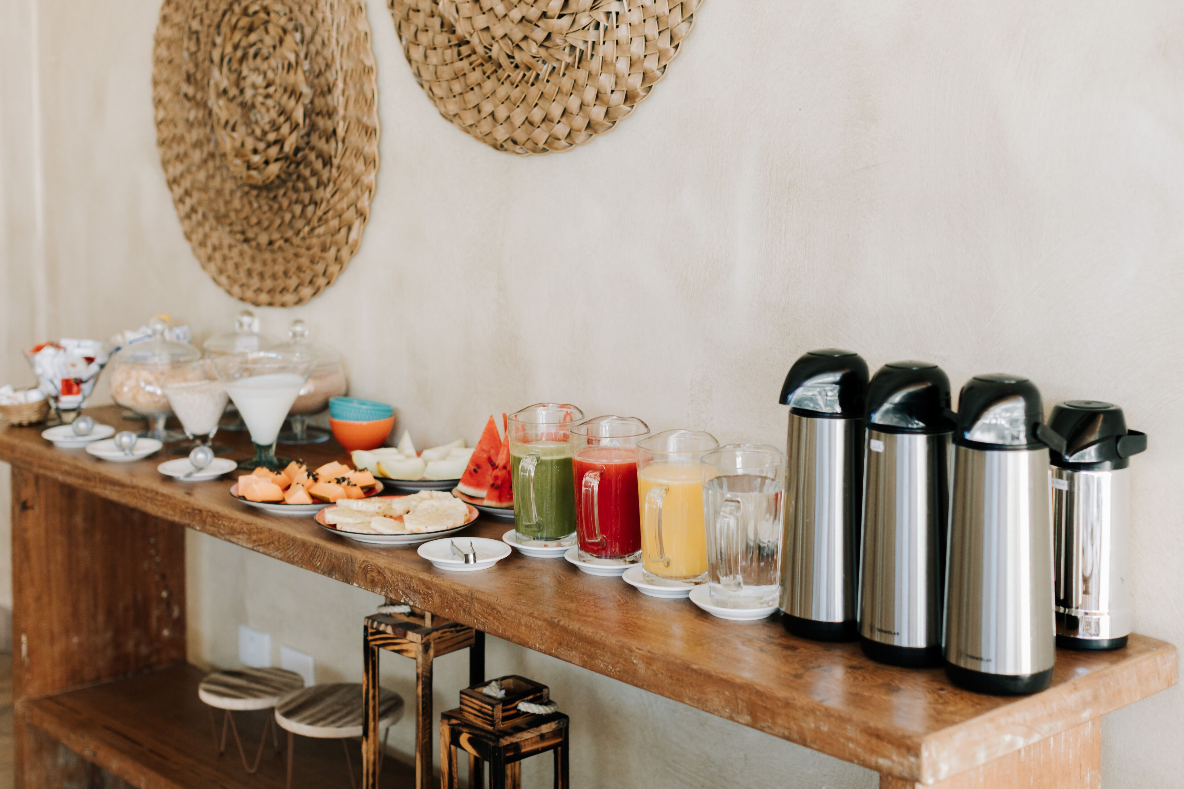 A breakfast buffet with various food items and beverages, including a selection of juices, a watermelon, and several thermos flasks.