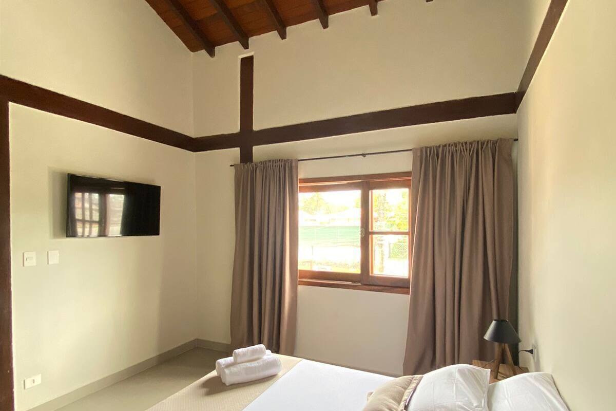 A bedroom with a wooden ceiling, a bed with white linens, a window with curtains, and a wall-mounted TV.