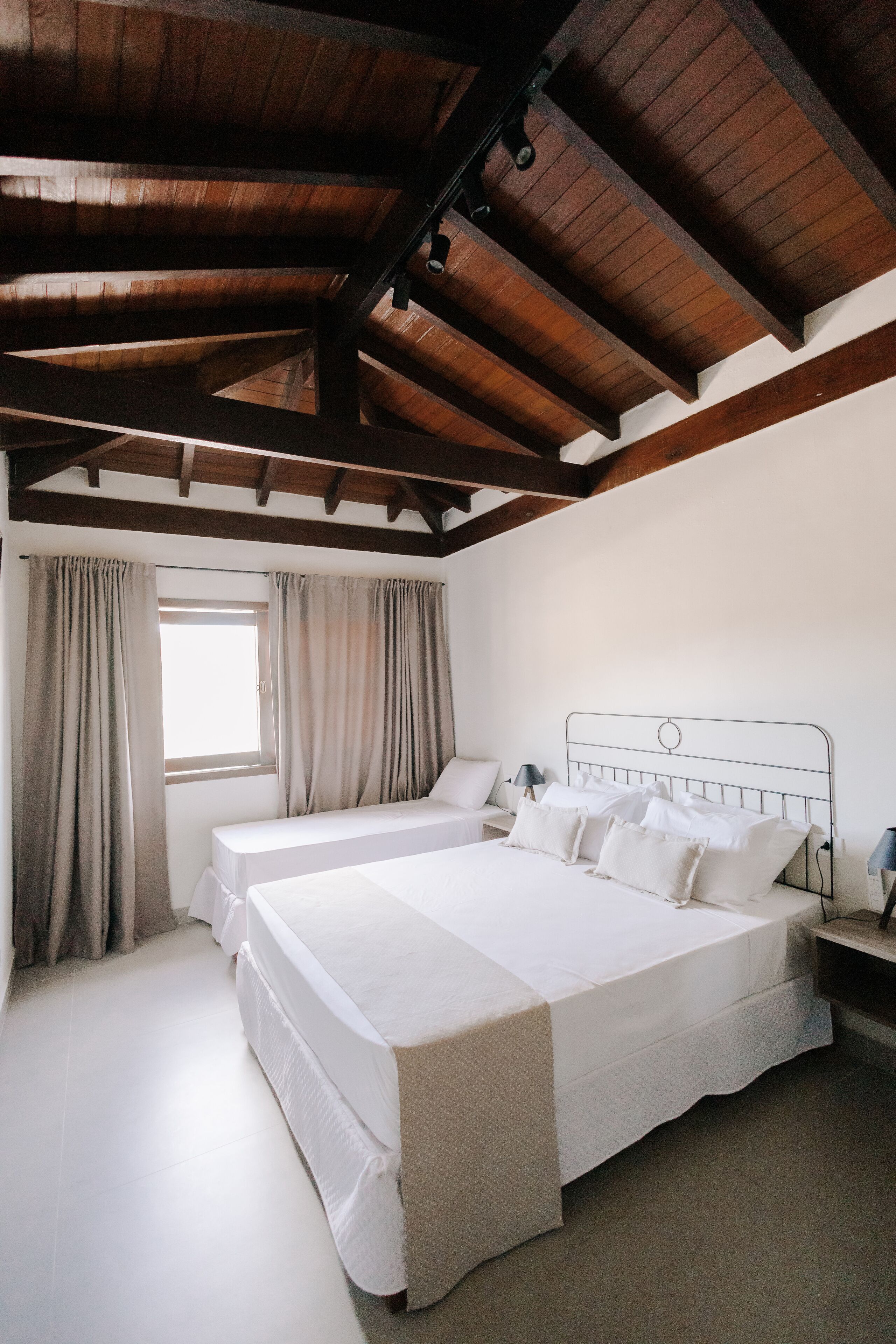 Quarto com uma cama grande, duas mesas de cabeceira, uma janela com cortinas e teto de madeira.
