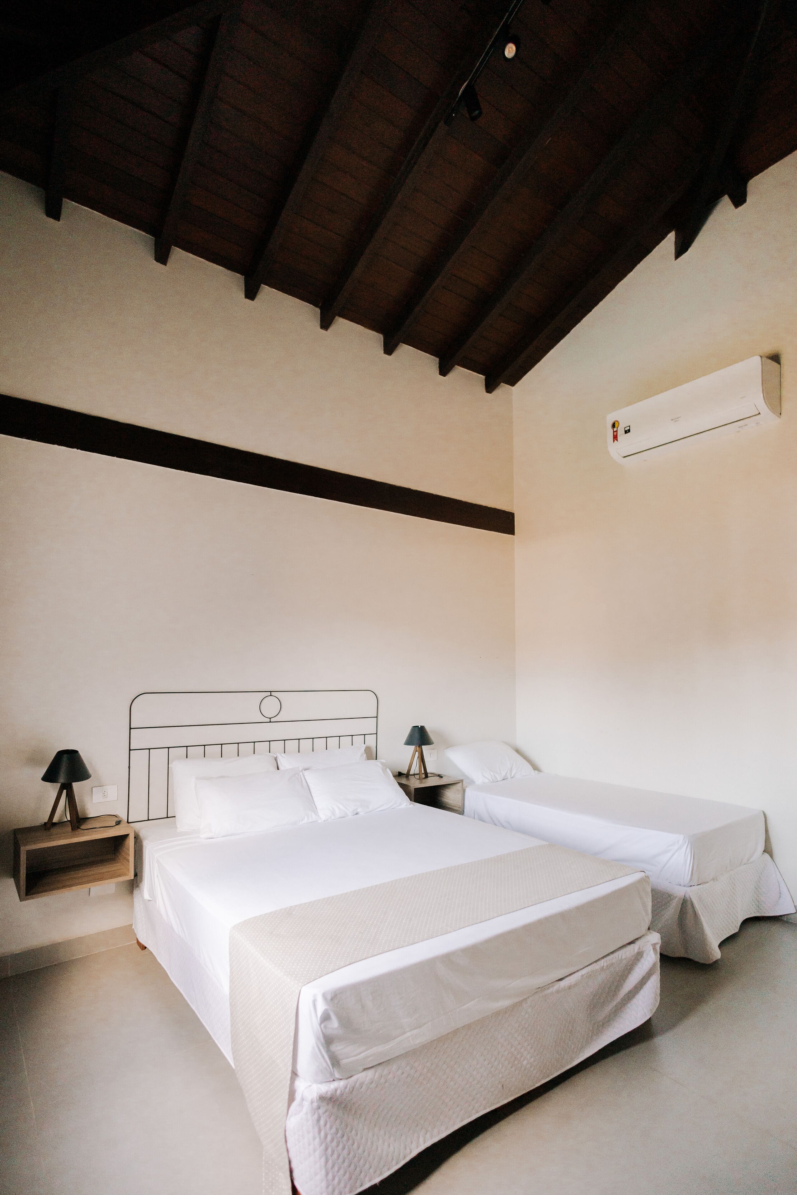 Une chambre avec deux lits, des poutres en bois au plafond, un climatiseur fixé au mur et des tables de chevet avec des lampes.