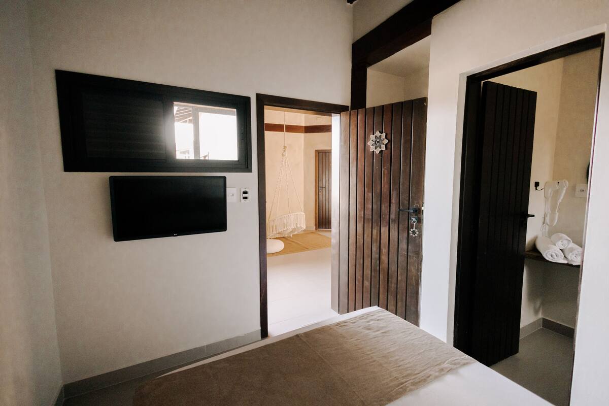 A hotel room with a bed, a flat-screen TV mounted on the wall, and a wooden door leading to a bathroom.