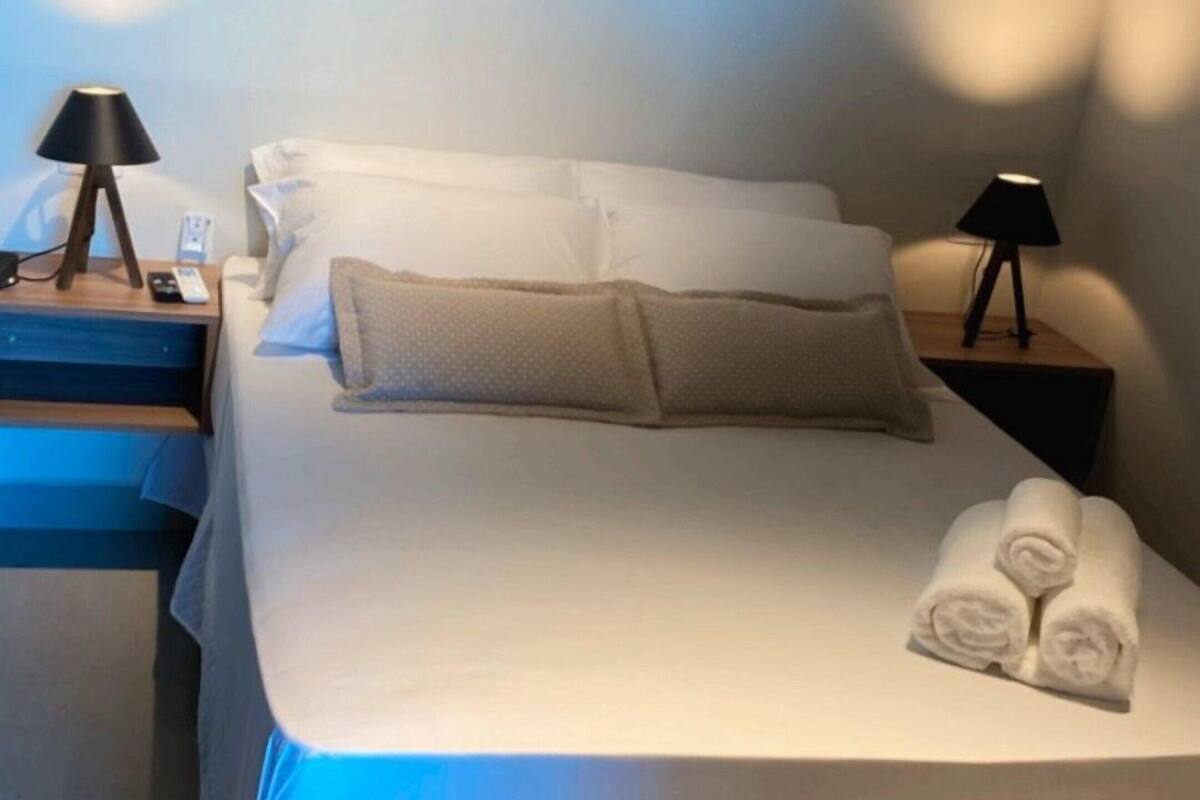 A neatly made bed with pillows, a rolled towel, and a bedside table with a lamp.