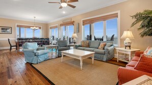 A spacious living room with a ceiling fan, a dining area with a table and chairs, and large windows offering a view. - Beach Buzz: Beachfront, Private Pool, Hot Tub (St. George Island)