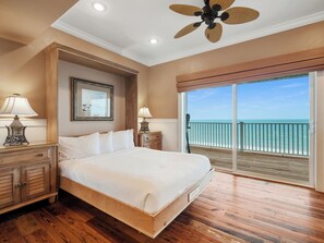 A bedroom with a large bed, wooden floor, ceiling fan, and a view of the ocean through a sliding door. - Beach Buzz: Beachfront, Private Pool, Hot Tub (St. George Island)
