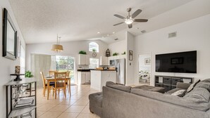 A spacious living room with a grey sofa, a dining area with wooden chairs, a kitchen with white cabinets, and a ceiling fan. - Beautiful 3-bedroom house, with private pool, backing onto golf course (Haines City)