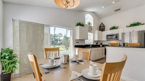 A modern kitchen with white cabinets, a microwave, and a sink. There is a dining area with a table set for two, and a large window offering a view of the outdoors. - Beautiful 3-bedroom house, with private pool, backing onto golf course (Haines City)