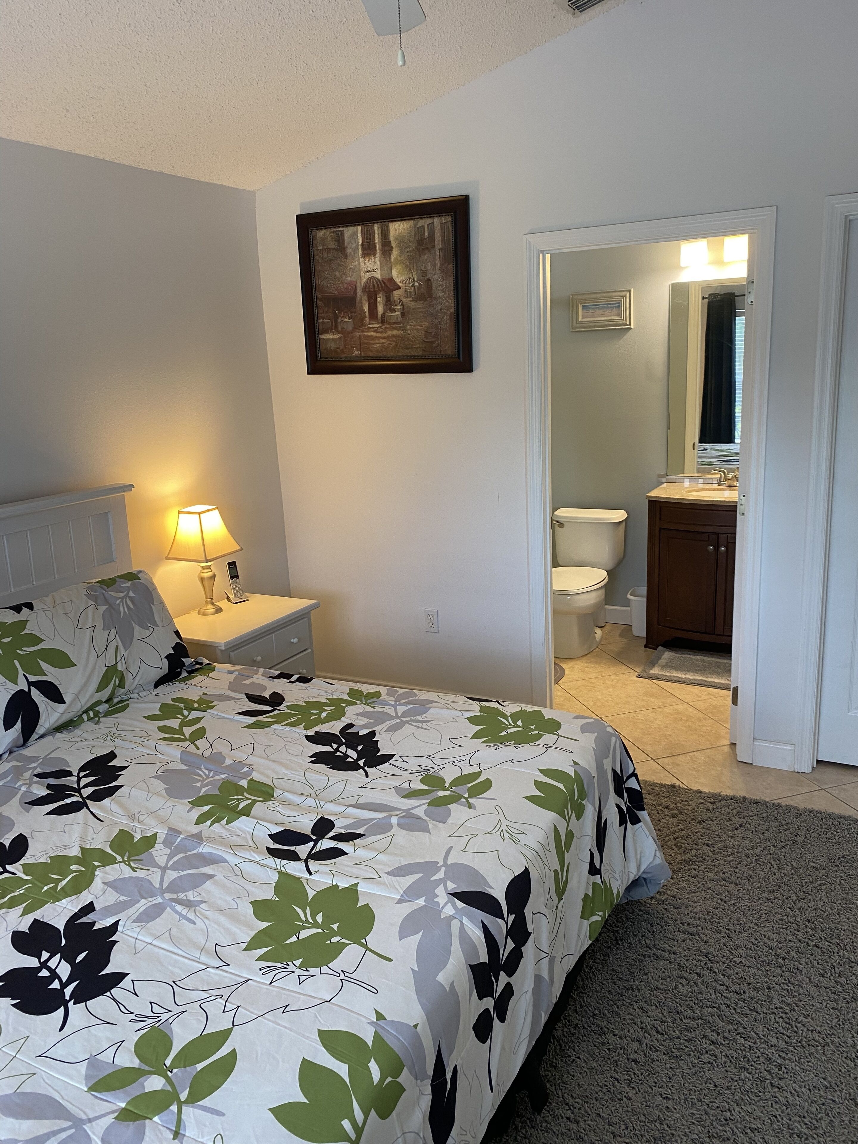 A bedroom with a bed, a nightstand, a lamp, a framed picture on the wall, and a bathroom visible through an open door.