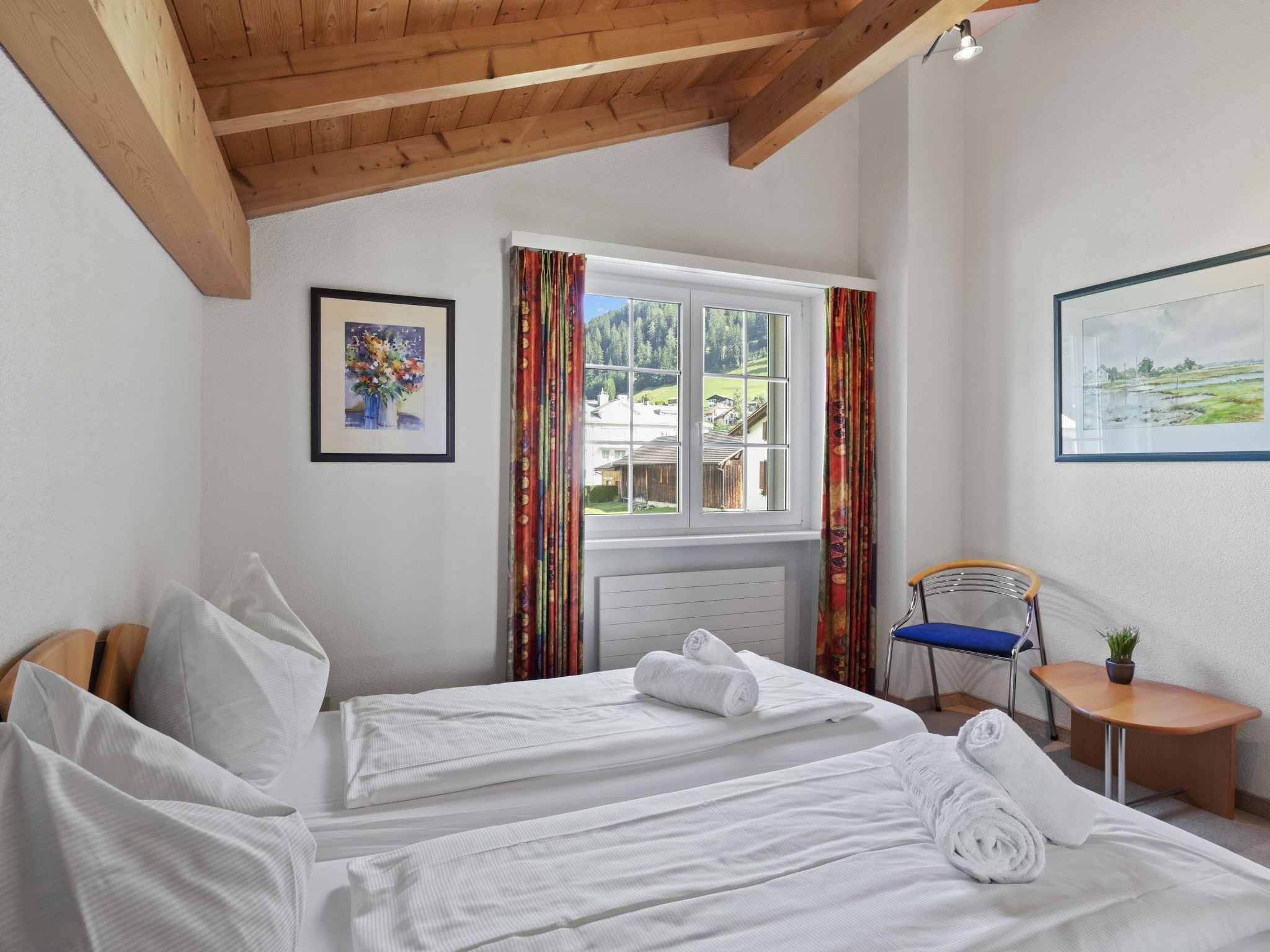 A hotel room with two beds, a window with curtains, a chair, a small table, and a painting on the wall.