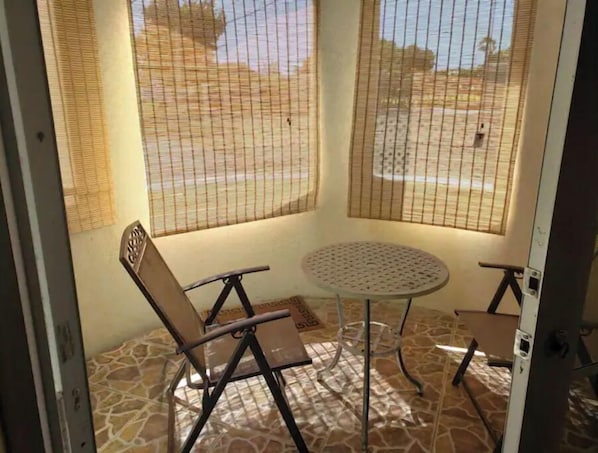 Een kamer met een klein rond tafeltje en twee stoelen, een raam met uitzicht op een strand en een betegelde vloer.
