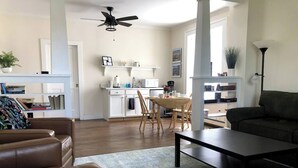 Amplia sala de estar con un sofá de cuero marrón, una mesa de centro oscura y una mesa de comedor con sillas. Incluye una zona de cocina con armarios blancos y un ventilador de techo.