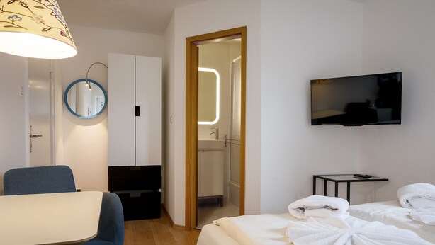 A hotel room with a bed, a desk, a chair, a TV, and a bathroom visible through an open door. - Greenhouse Apartments (Helgoland)
